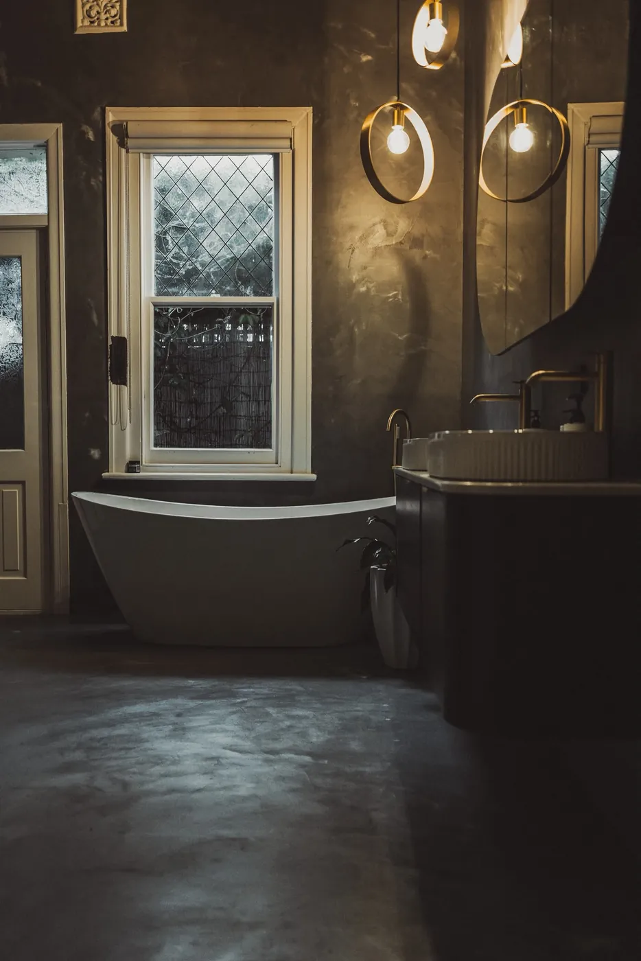 Bathroom with a dark microcement floor and walls, a freestanding bath and brass pendant lights