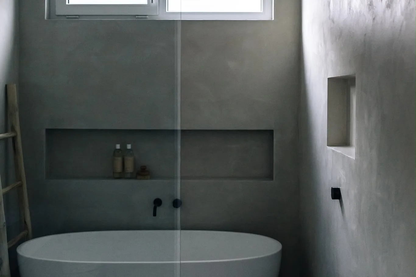 Microcement walls in low light around a freestanding bath and shower, with a long recessed shelf