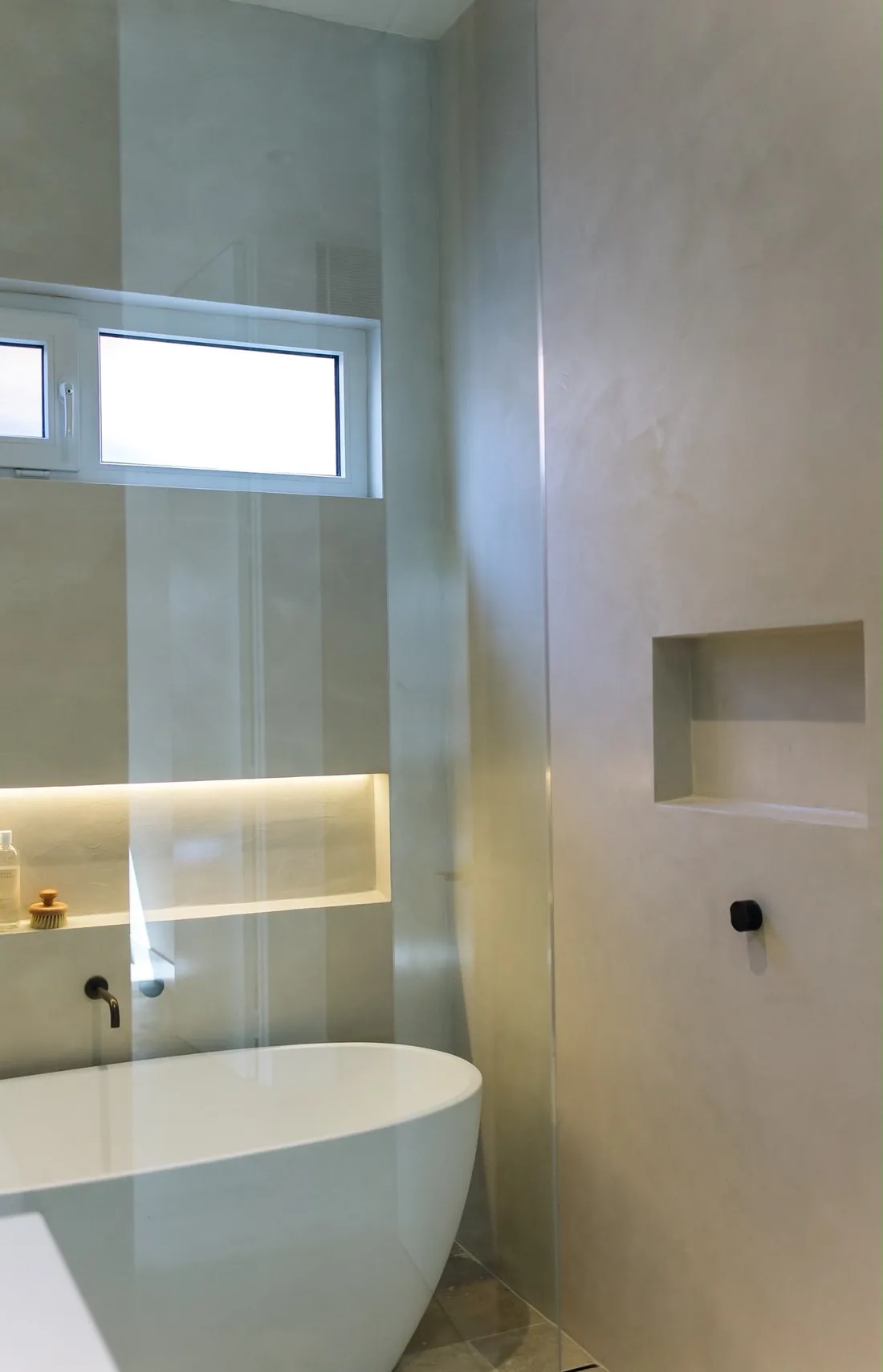 Microcement shower wall with a recessed niche beside a freestanding bath and a lit niche behind a glass screen