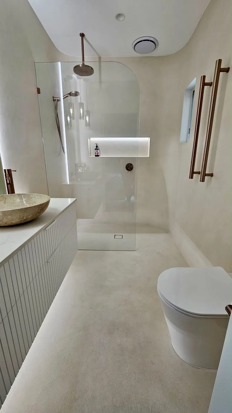 Bathroom with pale microcement walls and floor, a stone basin and a curved glass shower screen