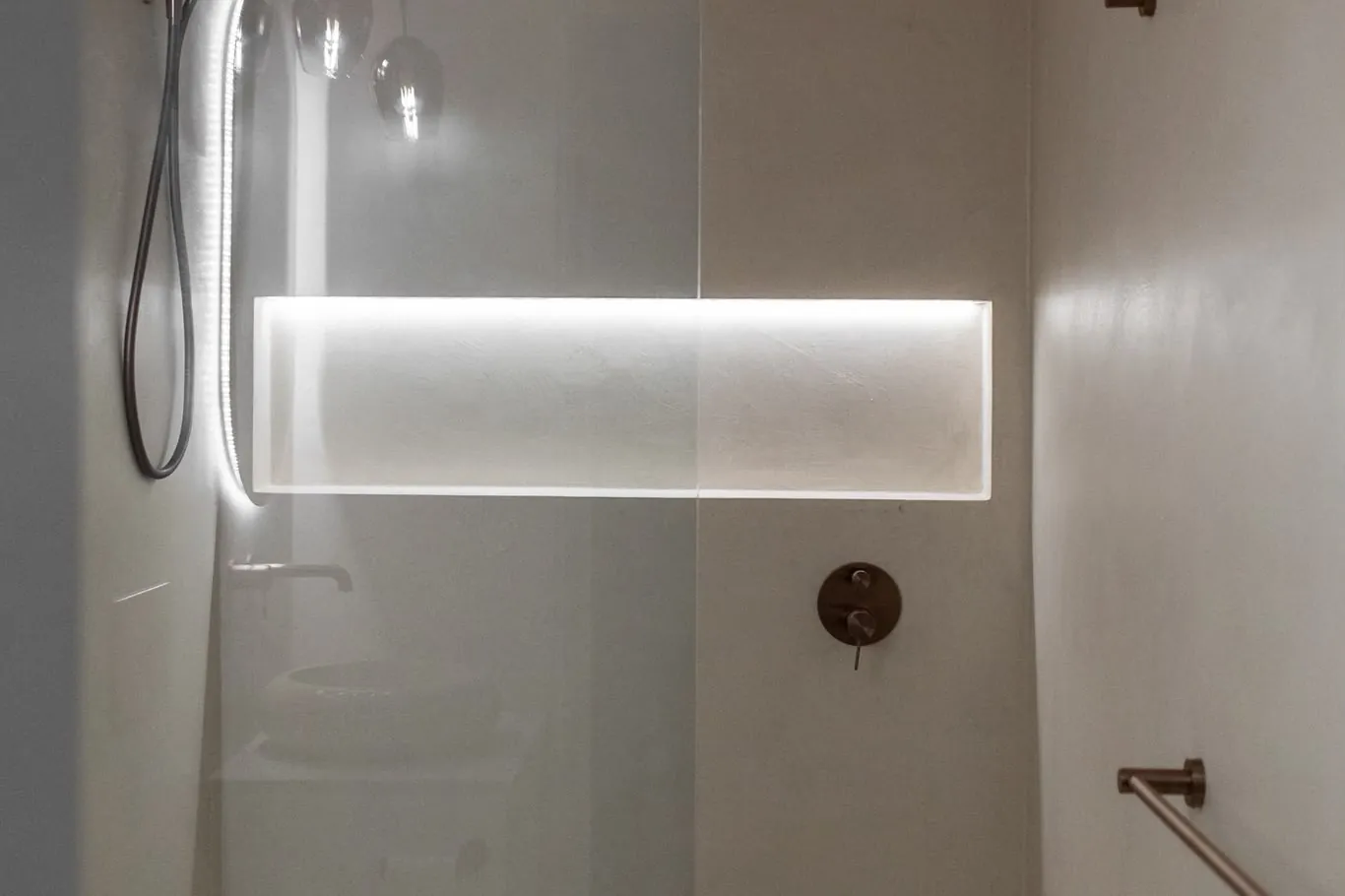 Shower with pale microcement walls, a long lit niche and bronze tapware behind a glass screen